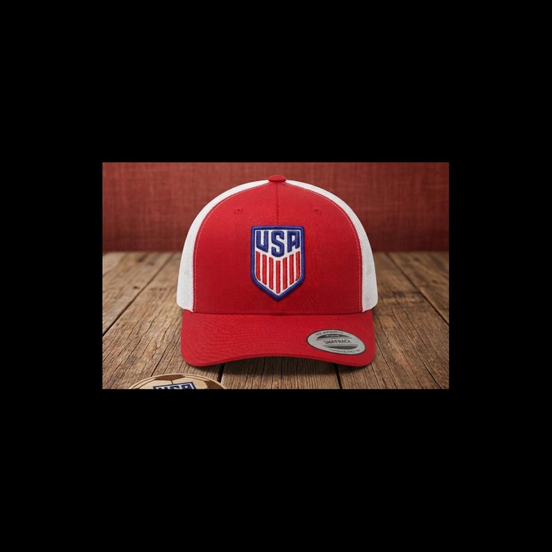 May include: A red and white baseball cap with a USA logo on the front. The cap has a curved brim and a mesh back. The USA logo is in a shield shape with red, white, and blue colors.