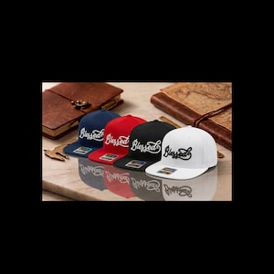May include: Four baseball caps in navy blue, red, black, and white, each with the word "Blessed" embroidered in white script. The caps are displayed on a reflective surface.