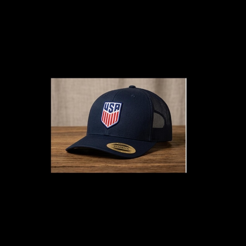 May include: A navy blue baseball cap with a mesh back and a curved brim. The front of the cap features a shield-shaped emblem with the letters "USA" in white and red stripes. The cap is sitting on a wooden surface.