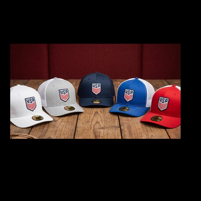 May include: Five baseball caps in white, gray, navy, blue, and red. Each cap features a white mesh back and a USA logo on the front. The caps are displayed on a wooden surface.