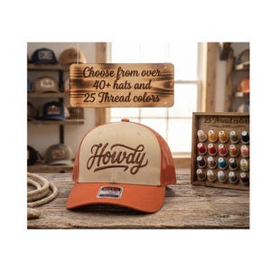 May include: A tan and orange trucker hat with the word Howdy embroidered in brown script on the front. A wooden sign above reads Choose from over 40+ hats and 25 Thread colors, and a wooden display rack shows 25 spools of various thread colors.