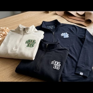 May include: Three quarter-zip pullovers in cream, black, and navy blue. Each garment features a stand-up collar, long sleeves, and an embroidered patch on the left chest displaying the text ALL 18 in a stylized font with a contrasting border.