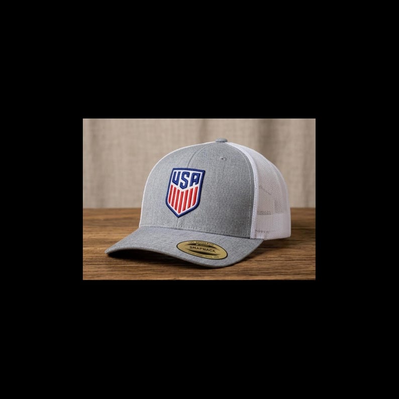 May include: A gray and white trucker hat with a USA shield logo. The front panel is gray with the USA logo in red, white, and blue. The mesh back is white, and the hat has a curved brim.
