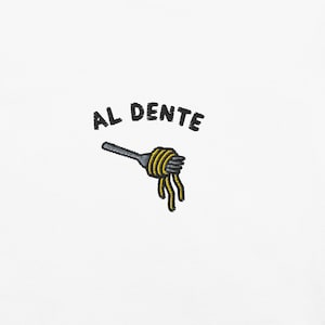 Puede incluir: Un diseño bordado sobre fondo blanco que muestra un tenedor gris enrollando espaguetis amarillos. Sobre el tenedor, el texto AL DENTE está bordado con hilo negro. El diseño está centrado y utiliza líneas sencillas para representar la pasta.