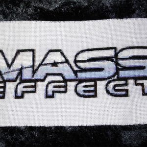 May include: Cross-stitch embroidery of the Mass Effect video game logo. The logo is white with a blue gradient and black outlines. The text "Mass Effect" is stitched in white with black outlines.