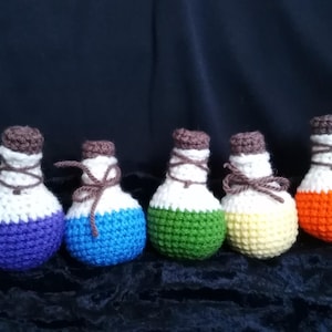 Könnte beinhalten: Sieben gehäkelte Zaubertrankflaschen in einem Regenbogen aus Farben, darunter Lila, Blau, Grün, Gelb, Orange und Rot. Jede Flasche hat einen weißen Deckel und einen braunen Korken.