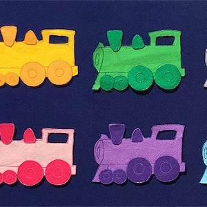 Puede incluir: Seis locomotoras de tren de juguete de fieltro en diferentes colores: amarillo, verde, gris, rosa, morado y azul. Cada tren tiene tres ruedas y una chimenea negra.