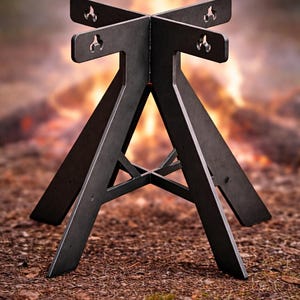 Könnte beinhalten: Ein dunkelgrauer Metall-Feuerstellenständer mit kreuzförmiger Oberseite und abgewinkelten Beinen. Der Ständer ist für eine Feuerstelle konzipiert und steht auf einem Bett aus braunen Blättern und Erde, mit einem verschwommenen Hintergrund eines Lagerfeuers.