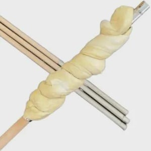 Op de afbeelding: Een close-up van lichtgekleurd, gedraaid deeg gewikkeld rond een houten deuvel en metalen staven. Het deeg lijkt ongebakken en heeft een zacht, getextureerd uiterlijk. De staven zijn in een kruisvorm gerangschikt.