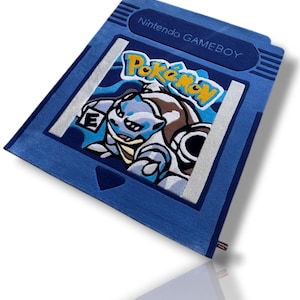 Könnte beinhalten: Blauer Teppich in Form einer Nintendo Gameboy-Cartridge mit dem Pokémon-Charakter Blastoise. Der Teppich zeigt das Wort "Pokemon" in Gelb und Orange sowie "Nintendo GAMEBOY" in Weiß.
