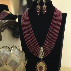 以下が含まれることがあります： マロン色のビーズネックレスと、ゴールドのペンダントとマッチするイヤリング。ペンダントには、小さなダイヤモンドに囲まれた大きなルビーの石が飾られています。