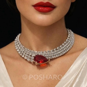 May include: A diamond necklace with a large, pear-shaped, red gemstone pendant. The necklace features multiple rows of small, clear diamonds. The image shows a close-up of the necklace on a person's neck.