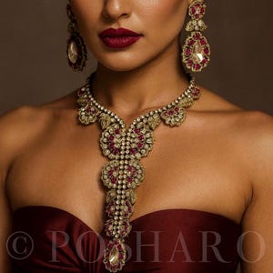 Puede incluir: Un collar dorado y pendientes a juego con piedras preciosas de color rojo rubí y transparentes. El collar presenta un diseño de colgante central y se combina con un top sin tirantes burdeos. Los pendientes son de estilo colgante.