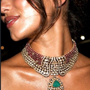 May include: A close-up of a luxurious necklace. The necklace features a wide collar with alternating rows of red stones and clear crystals. A large, ornate pendant with a green gemstone hangs from the center, showcasing intricate detailing.