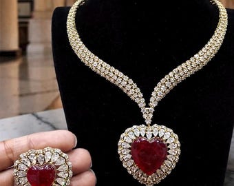 Op de afbeelding: Een hartvormige robijn hangerketting en bijpassende ring. De ketting en ring hebben een grote, hartvormige rode steen omringd door heldere, sprankelende stenen gezet in een goudkleurig metaal. De ketting wordt getoond op een zwarte mannequin.