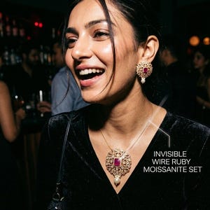 Może przedstawiać: Kobieta nosi naszyjnik i kolczyki "INVISIBLE WIRE RUBY MOISSANITE SET". Biżuteria posiada duży, centralny rubin otoczony mniejszymi, przezroczystymi kamieniami. Kolczyki mają podobny design z perłową kroplą.