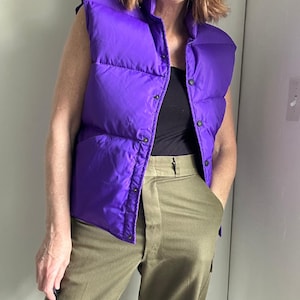 May include: A vibrant purple puffer vest with a high collar and snap closures. The vest is worn over a black top and paired with olive green pants. The vest's quilted design adds texture, creating a bold and stylish look.