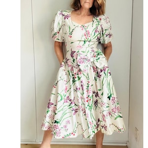 Może przedstawiać: Biała sukienka w stylu vintage z kwiatowym wzorem z fioletowymi i różowymi kwiatami oraz zielonymi liśćmi. Sukienka ma krótkie, bufiaste rękawy, kwadratowy dekolt i dużą kokardę w talii. Spódnica sięga do kolan.