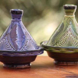 May include: Two ceramic tagines with conical lids, one in deep purple and one in olive green. Both feature a wide, shallow base and a narrow top knob, decorated with intricate, hand-carved radial patterns and embossed lines around the rim.