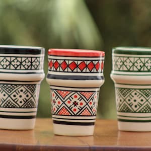 May include: Three ceramic cups with a tapered shape and a flared rim. The left cup features black geometric patterns, the center cup has red and black designs, and the right cup displays green geometric motifs on a white background.