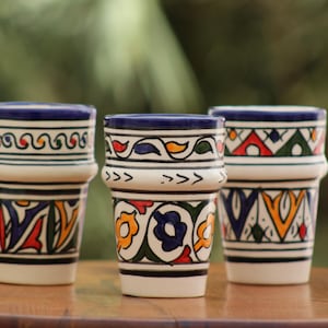 May include: Three ceramic cups featuring hand-painted designs in blue, yellow, green, and red. Each cup has a distinct pattern of geometric shapes, floral motifs, and wavy lines, with a prominent horizontal ridge encircling the upper section.