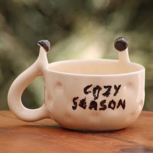 Puede incluir: Una taza de cerámica color crema con una superficie con hoyuelos y el texto "COZY SEASON" escrito en letras marrón oscuro. La taza presenta un asa con forma de cuerpo de caracol con dos tallos oculares de color marrón oscuro en el borde.
