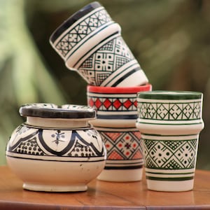May include: A collection of ceramic items including a rounded bowl with a black painted pattern and three cylindrical cups. The cups feature white bases with intricate geometric designs in black, red, and green paint, stacked and arranged together.