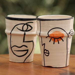 May include: Two off-white ceramic cups featuring black line art of abstract faces. The cup on the left shows a profile with a large eye and lips, while the cup on the right displays a closed eye with an orange painted eyelid and black lips.