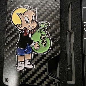 Puede incluir: Una cartera de fibra de carbono negra con un emblema de esmalte colorido de un niño de dibujos animados con cabello rubio, pajarita roja y pantalones cortos azules sosteniendo una bolsa de dinero verde con un signo de dólar. Incluye una herramienta.