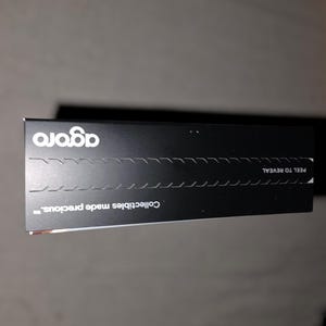 May include: A black rectangular box with the word "agora" in white, stylized font. The box has a perforated line for peeling and the text "Collectibles made precious."