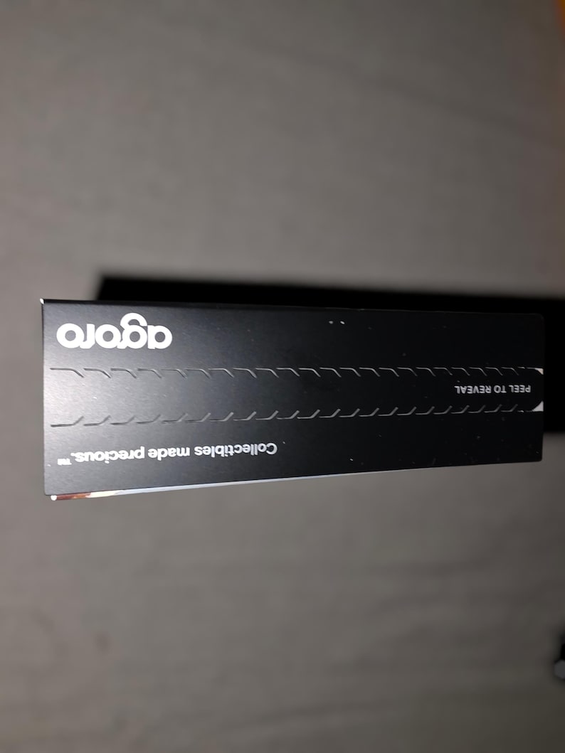 May include: A black rectangular box with the word "agora" in white, stylized font. The box has a perforated line for peeling and the text "Collectibles made precious."