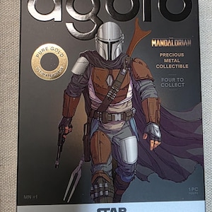 May include: A black box featuring a detailed illustration of The Mandalorian from Star Wars. The box includes text that reads "agoro," "Pure Gold," "Precious Metal Collectible," and "Engraved Foil Blind Box."