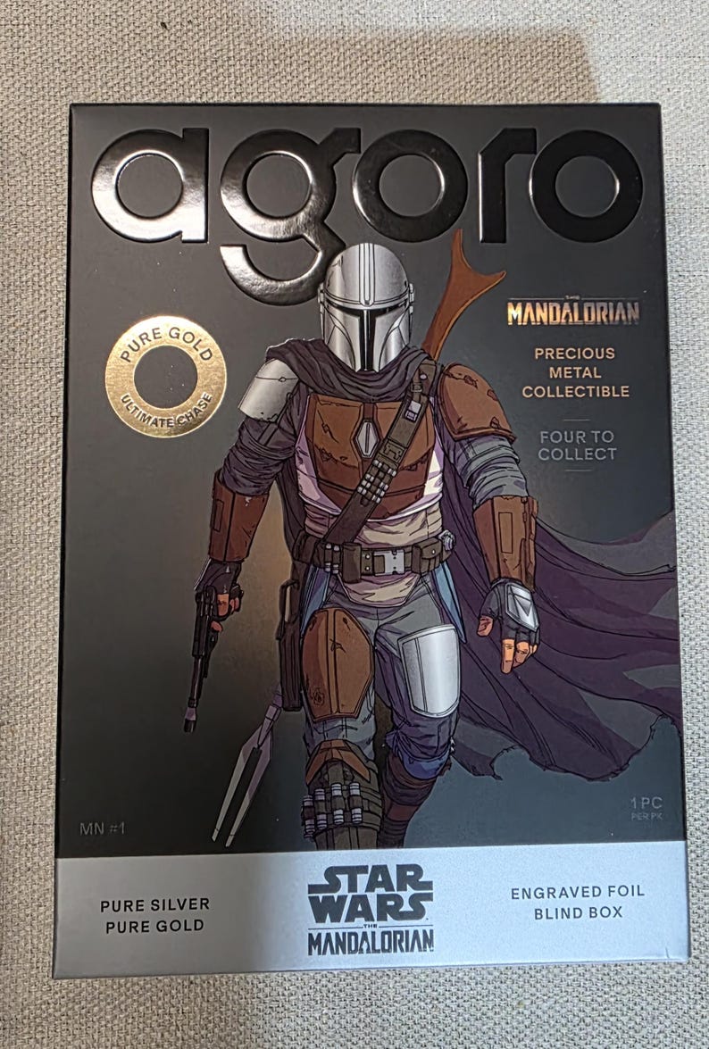 May include: A black box featuring a detailed illustration of The Mandalorian from Star Wars. The box includes text that reads "agoro," "Pure Gold," "Precious Metal Collectible," and "Engraved Foil Blind Box."
