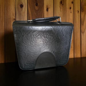 May include: Black leather briefcase with a handle and silver-colored metal hardware. The briefcase has a textured surface and a curved, black accent at the bottom. The background is a wooden wall.