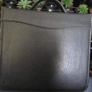 May include: A black leather briefcase with a top handle. The briefcase has a textured surface and a curved design element near the top. A small logo is visible in the lower left corner.