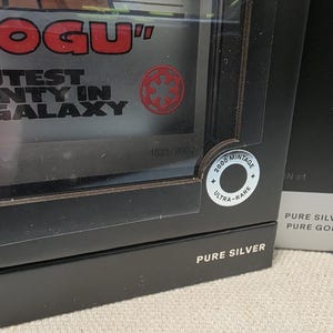 May include: A black box with the words "ROGU" and "CUTEST COUNTY IN THE GALAXY" in red and white. A circular sticker reads "2000 MINTAGE ULTRA-RARE". The box is labeled "PURE SILVER".
