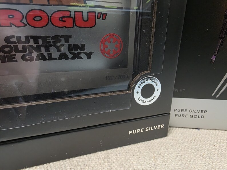 May include: A black box with the words "ROGU" and "CUTEST COUNTY IN THE GALAXY" in red and white. A circular sticker reads "2000 MINTAGE ULTRA-RARE". The box is labeled "PURE SILVER".