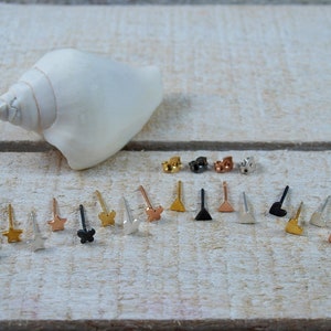 May include: A selection of small, minimalist earrings in gold, silver, black, and rose gold. The earrings feature various shapes, including stars, hearts, triangles, and a butterfly.