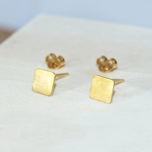 Puede incluir: Un par de pendientes de botón cuadrados de oro. Los pendientes tienen un acabado cepillado y son simples y elegantes.