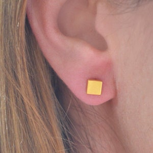 May include: A pair of small, square-shaped gold earrings. The earrings are worn in the earlobes of a person with long brown hair.