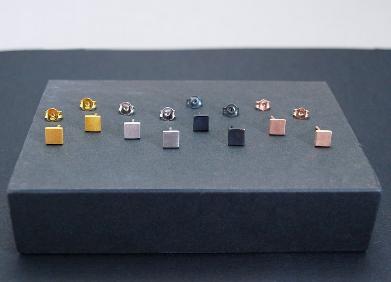 May include: A set of 10 square stud earrings in various colors, including gold, silver, black, and rose gold. The earrings are displayed on a black surface.