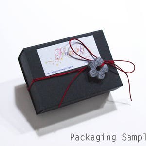 May include: A black gift box with a red ribbon and a gray felt flower. The box has a label with the text "Fraulein" and a website address.