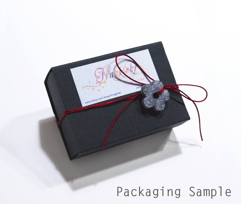 May include: A black gift box with a red ribbon and a gray felt flower. The box has a label with the text "Fraulein" and a website address.