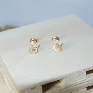 May include: A pair of gold-toned stud earrings with a circular design. The earrings are on a white wooden surface.