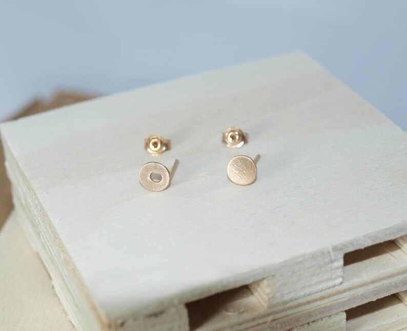 May include: A pair of gold-toned stud earrings with a circular design. The earrings are on a white wooden surface.