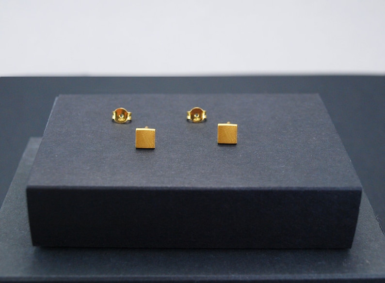 May include: Three gold square stud earrings on a black display box.