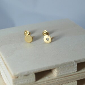 May include: A pair of gold-toned stud earrings with a circular design. The earrings are on a light wood surface.