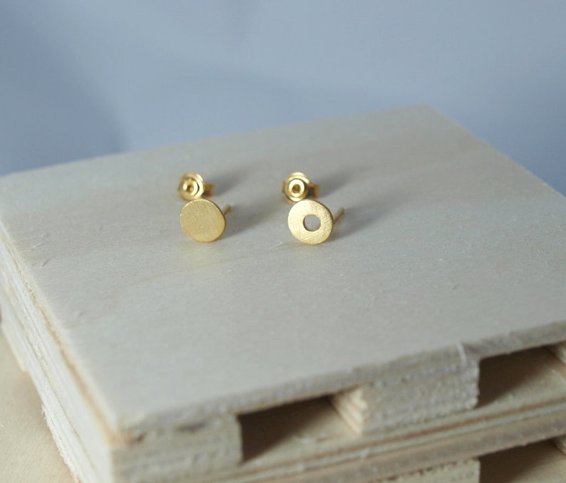 May include: A pair of gold-toned stud earrings with a circular design. The earrings are on a light wood surface.