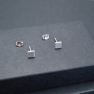 May include: Three silver square stud earrings on a gray surface. The earrings are small and simple.