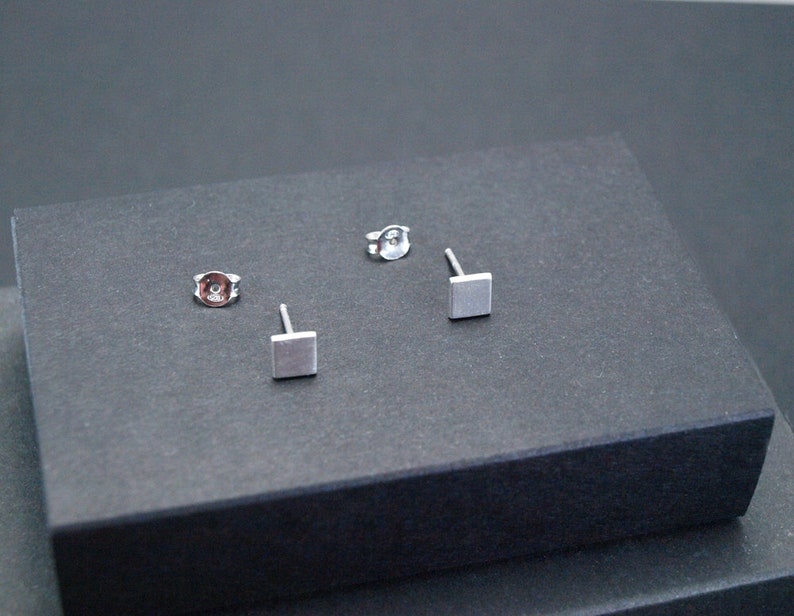 May include: Three silver square stud earrings on a gray surface. The earrings are small and simple.